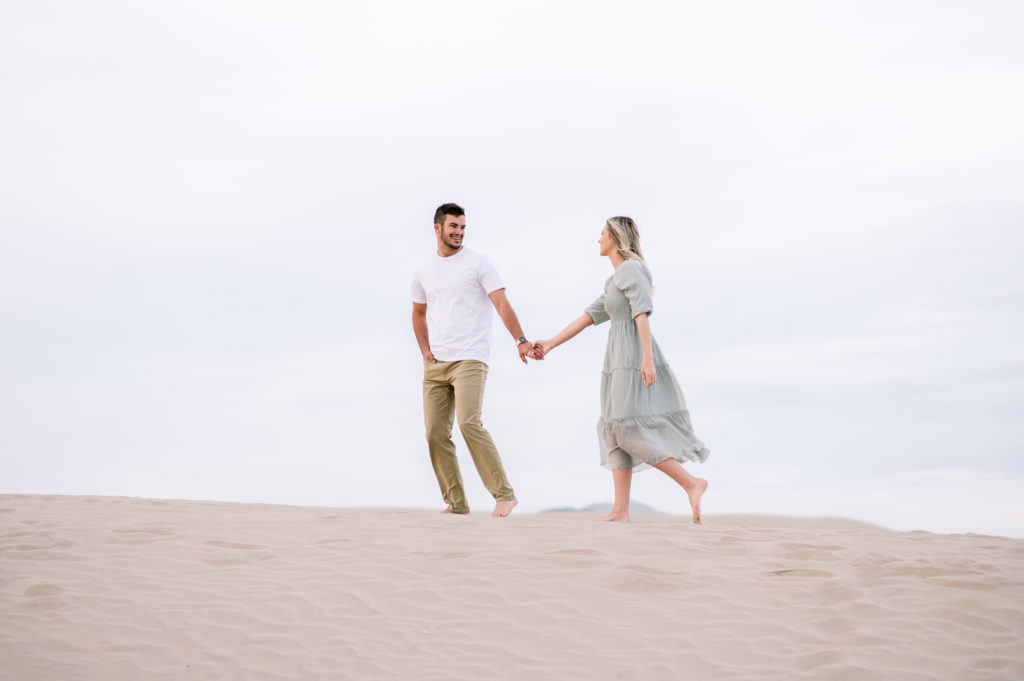 Cute engagement picture taken by Andria Joleen Photography from a couples session at the Little Sahara Sand Dunes. The couple is holding hands and walking across the top of one of the sand dunes. 