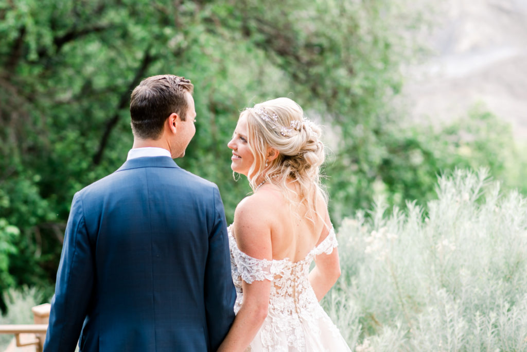 Romantic couple walking away from the camera looking at each other and smiling on their wedding day. They are walking down a path at Utah wedding venue Louland Falls on the way to their reception. She is wearing an off the shoulder lace dress and he is in a blue suit. 